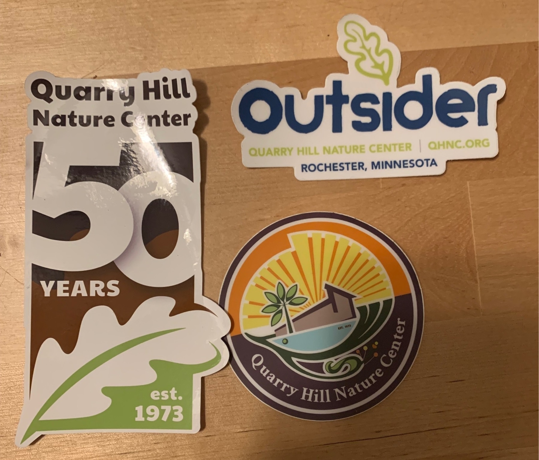 3 stickers. Quarry Hill Nature Center 50 years with a leaf. Outsider Quarry Hill Nature Center. Quarry Hill Nature Center stylized with a sun behind and the pond in front on a round sticker. 